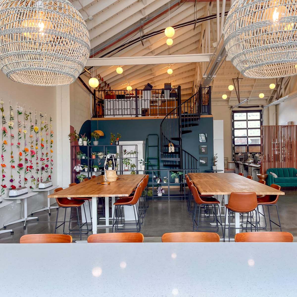 A spacious interior featuring wooden tables, woven pendant lights, and a spiral staircase, with a separation of seating areas and decorative plants on the walls.