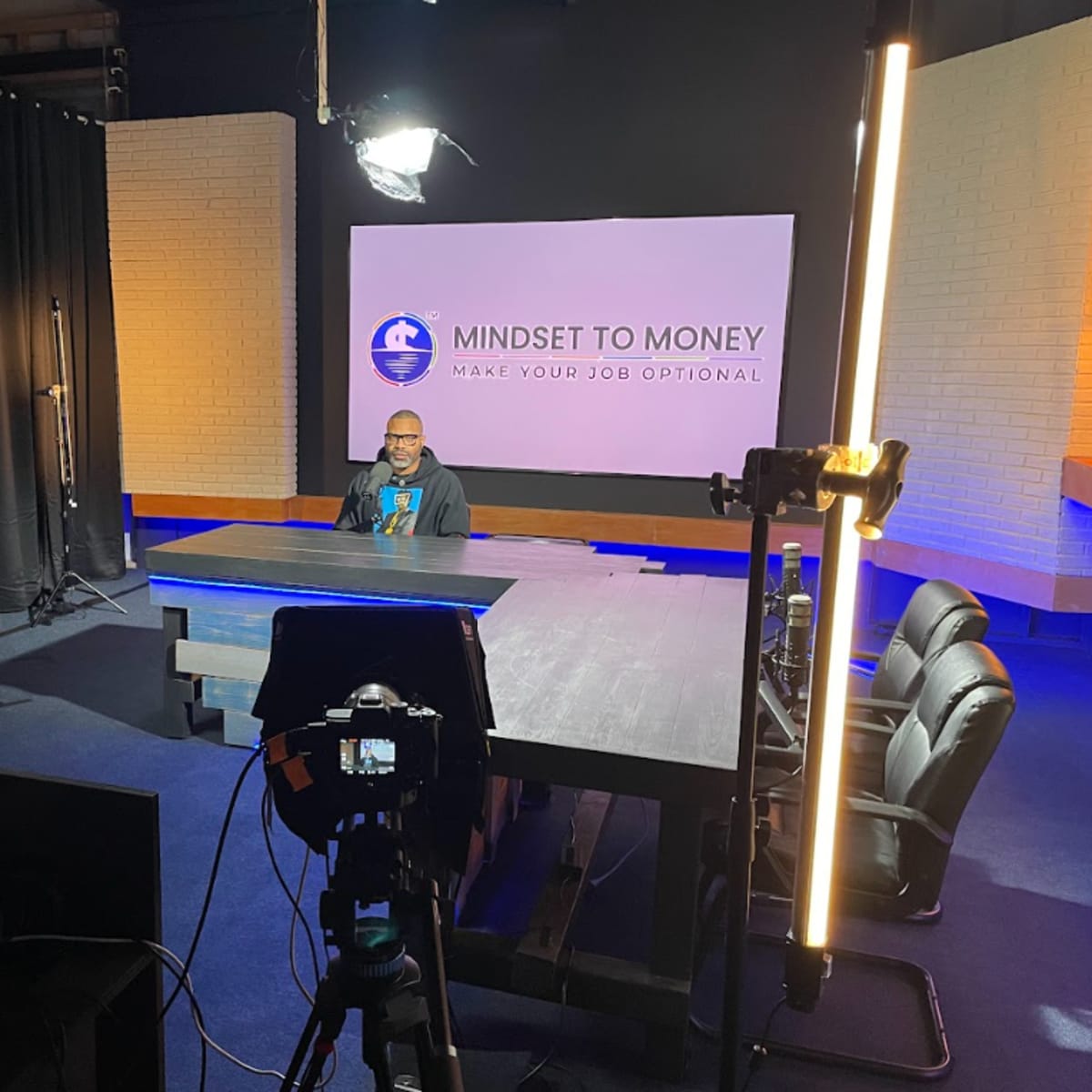 A speaker sits at a large table in a well-lit studio, with cameras and equipment set up for a production labeled "Mindset to Money."