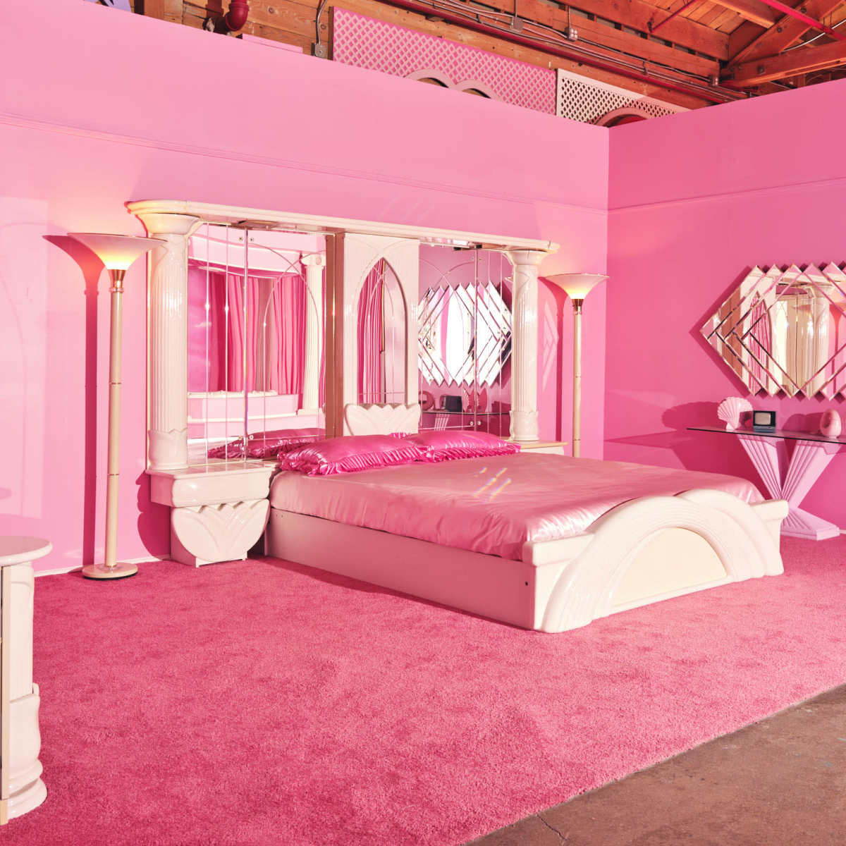 A pink-themed bedroom featuring a bed with a pink comforter, matching walls, and mirrored furniture.
