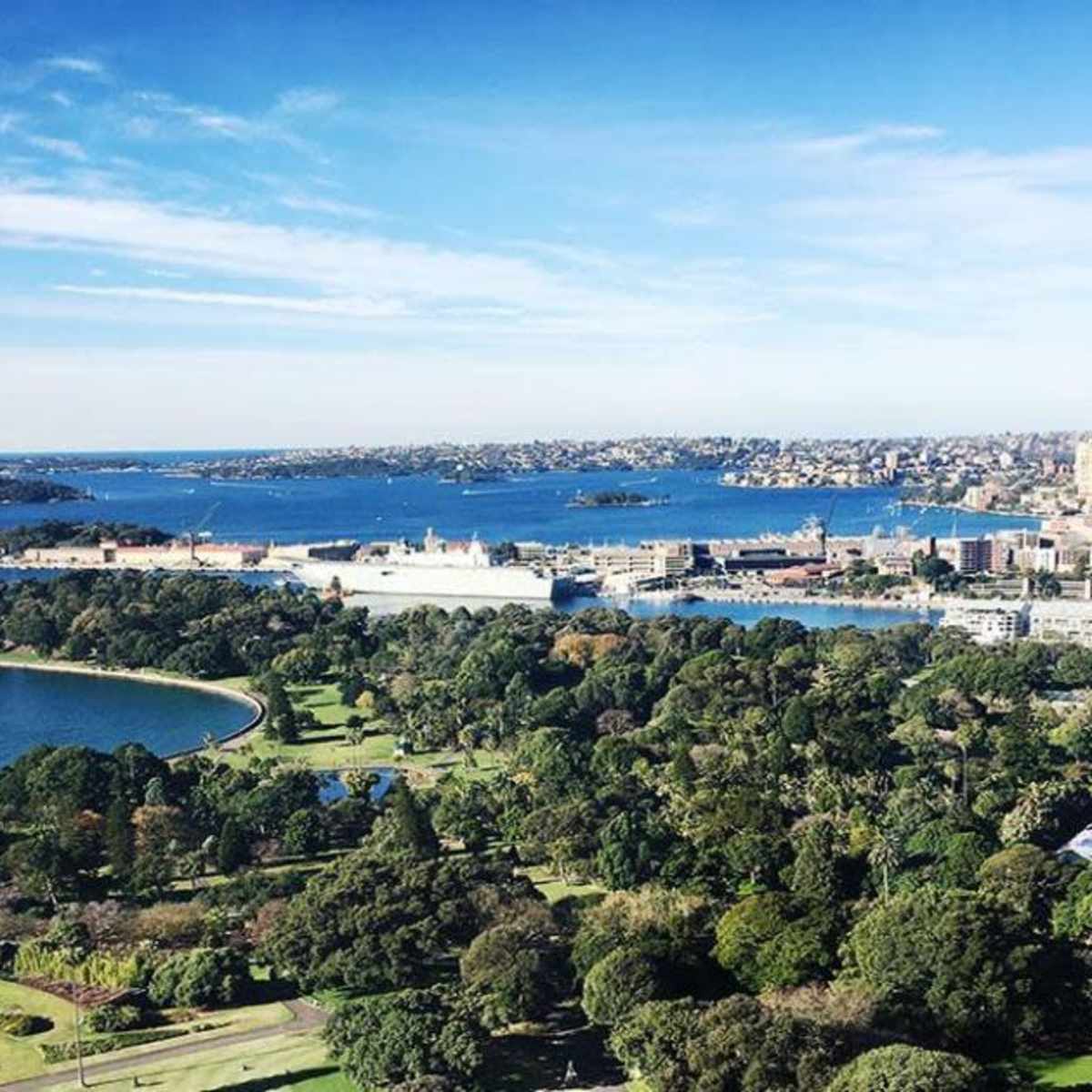 A panoramic view of a coastal city with lush green parks, a harbor, and surrounding waterways under a clear blue sky.