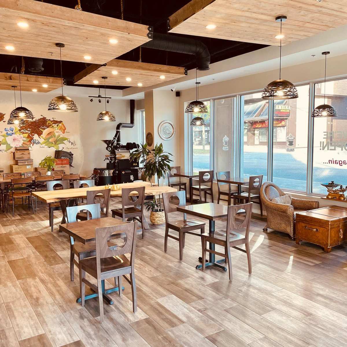 A spacious café with wooden tables and chairs, overhead pendant lights, and large windows allowing natural light to enter.