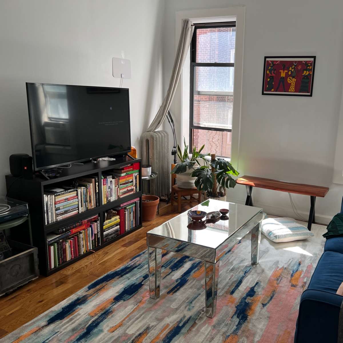 A living room features a blue sofa, a coffee table, a TV unit filled with books, and plants, with sunlight streaming through the windows.