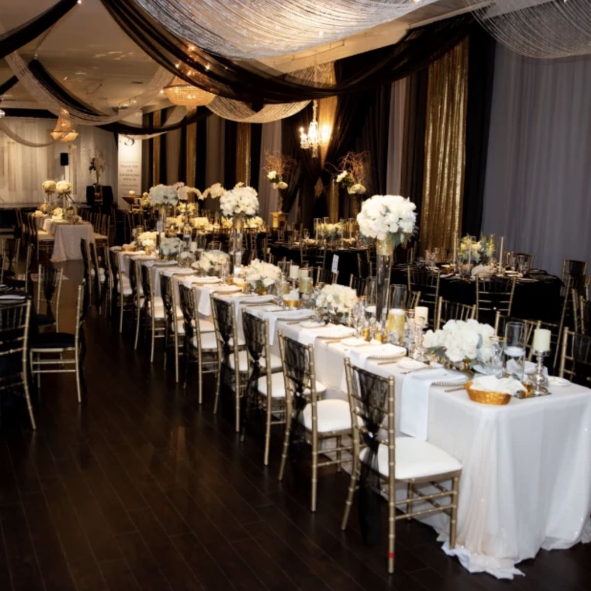 An elegantly arranged banquet hall with long tables set for a formal event, decorated with white flowers and black and gold drapery.