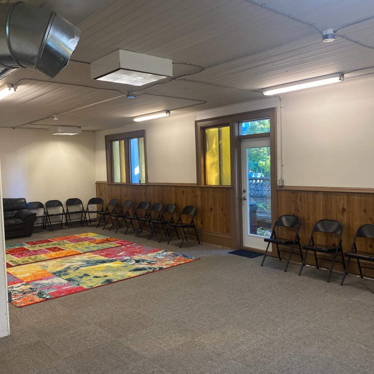 An empty room with a patterned rug in the center and rows of folding chairs along the walls, illuminated by overhead lights.