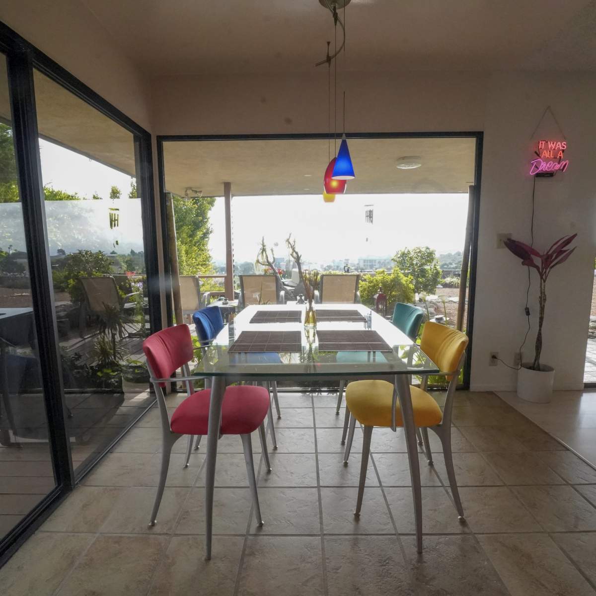 A glass dining table with colorful chairs is set against large windows showcasing a garden and outdoor seating area.