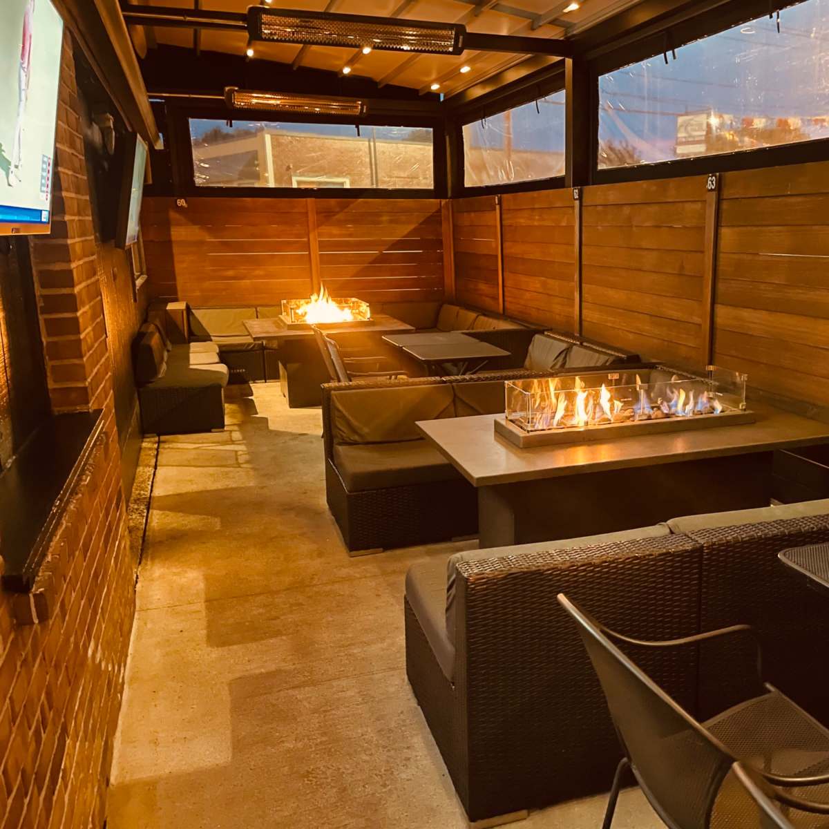 A cozy outdoor seating area with dark wicker furniture and a gas fire pit in the center, enclosed by wooden walls and a roof.
