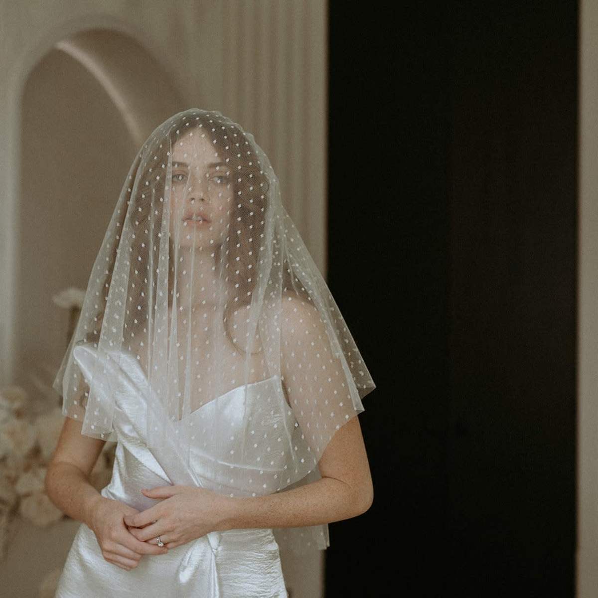 A bride wearing a polka-dot veil stands elegantly in a softly lit indoor setting.