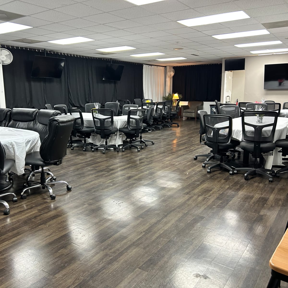 A spacious meeting room with several tables covered in white tablecloths and black rolling chairs arranged around them.