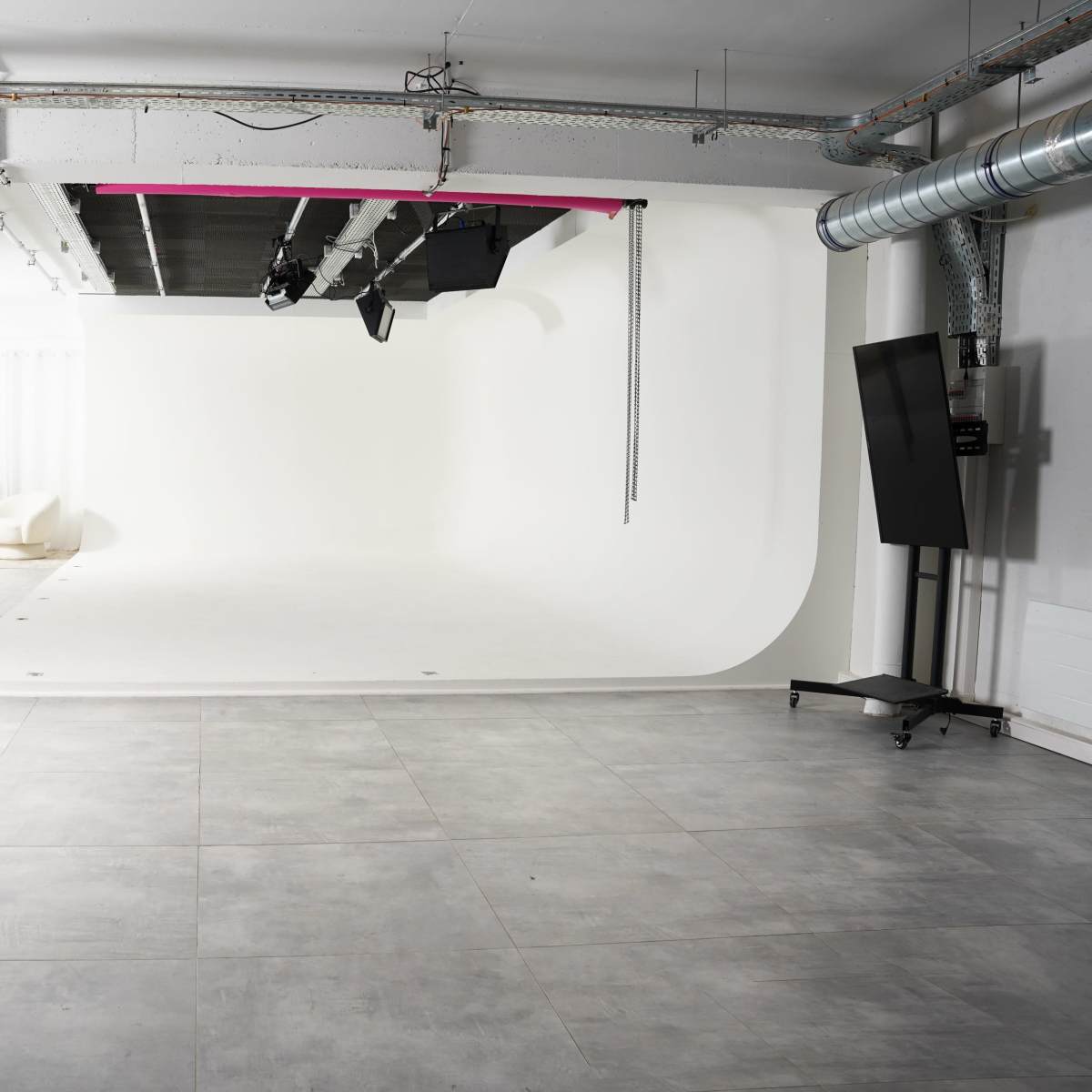 A spacious photography studio with a seamless white backdrop and a light setup, featuring grey tiled flooring and modern furniture in the corner.
