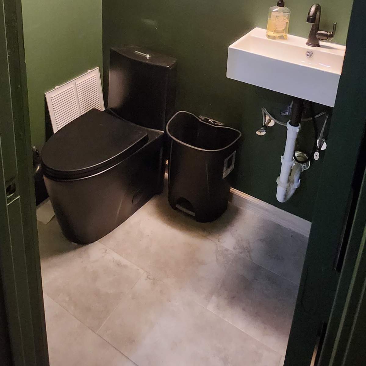 A small bathroom with dark green walls, featuring a black toilet, a sink with a modern faucet, and a trash can.