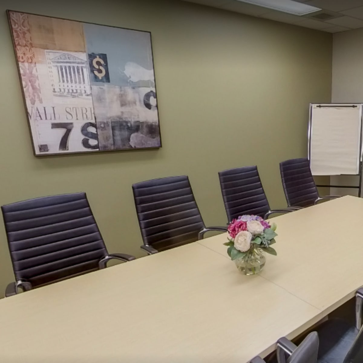 A conference room features a long table surrounded by several black chairs, a flower arrangement in the center, and a large screen mounted on the wall.