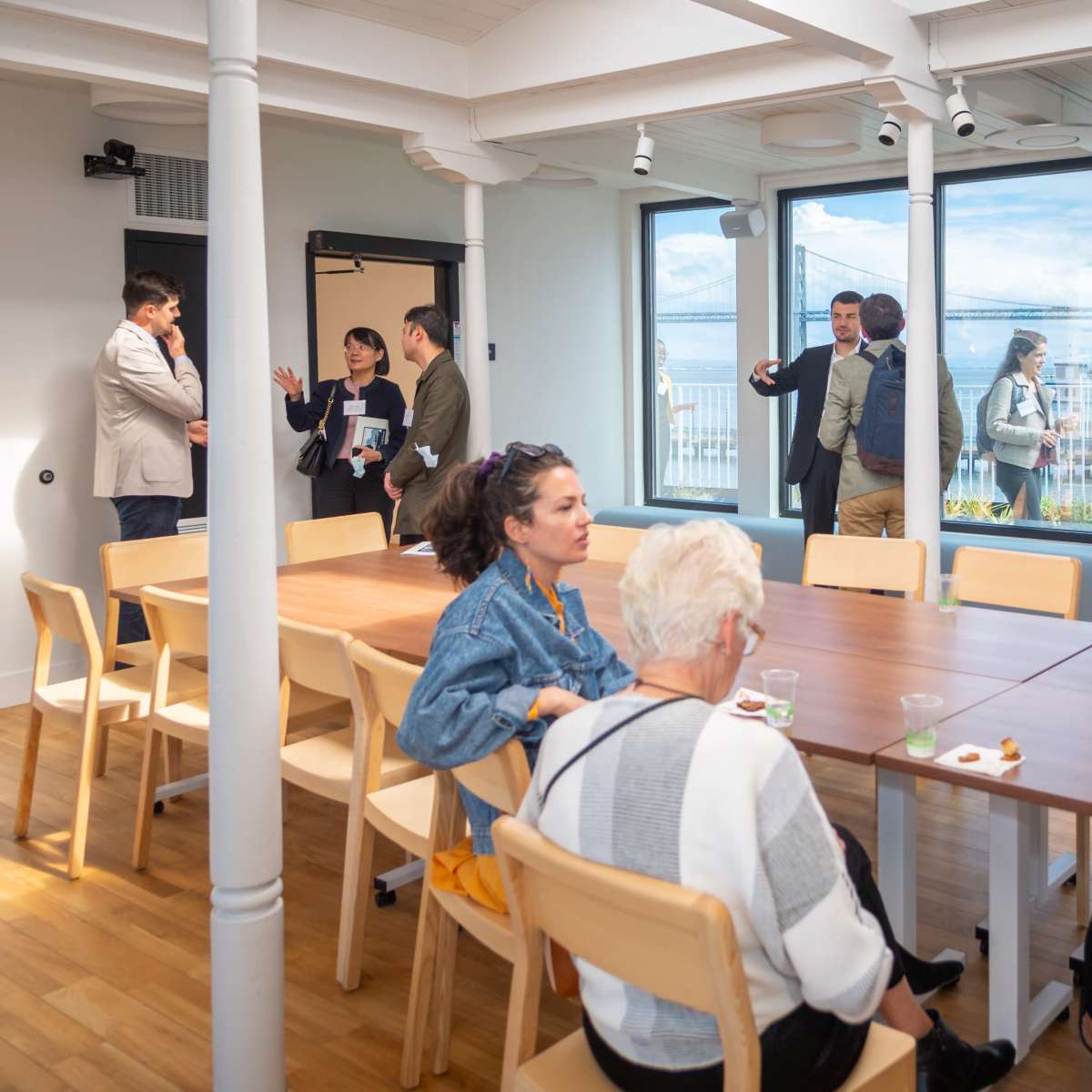 A group of people converses around a long table in a well-lit room with large windows overlooking a cityscape.