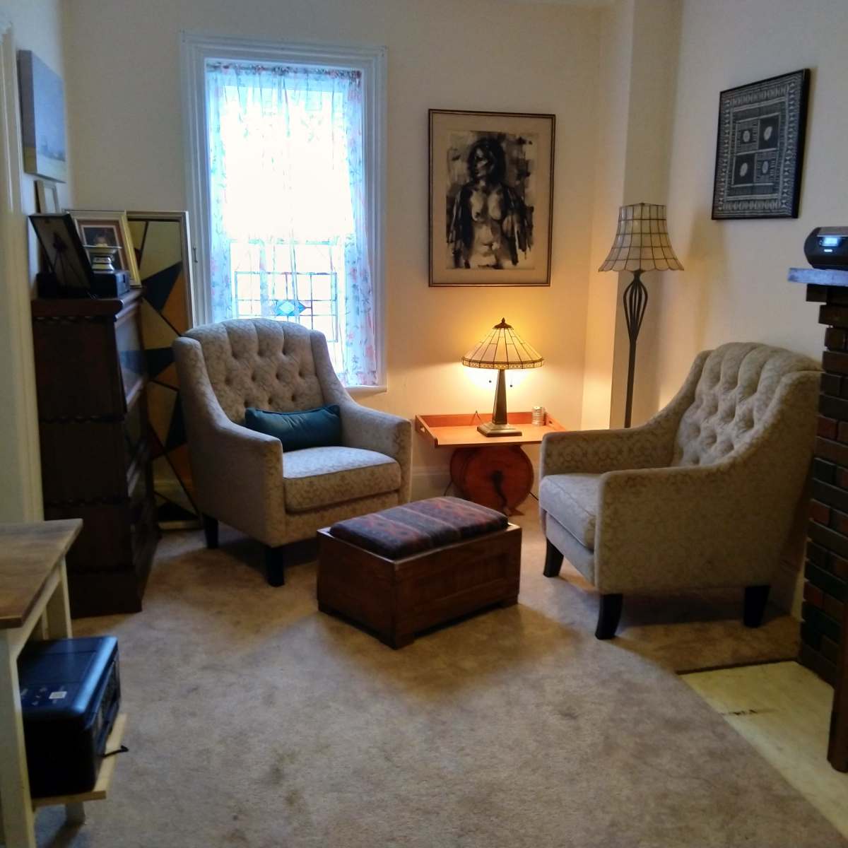 A cozy living room with two upholstered chairs, a small wooden table with a lamp, and a brick fireplace.