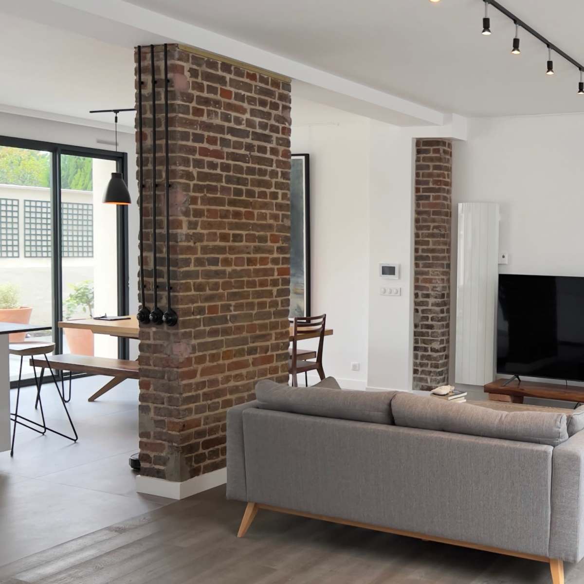 A modern living room with a gray sofa, a flat-screen TV on a wooden stand, exposed brick walls, and a potted plant in the corner.
