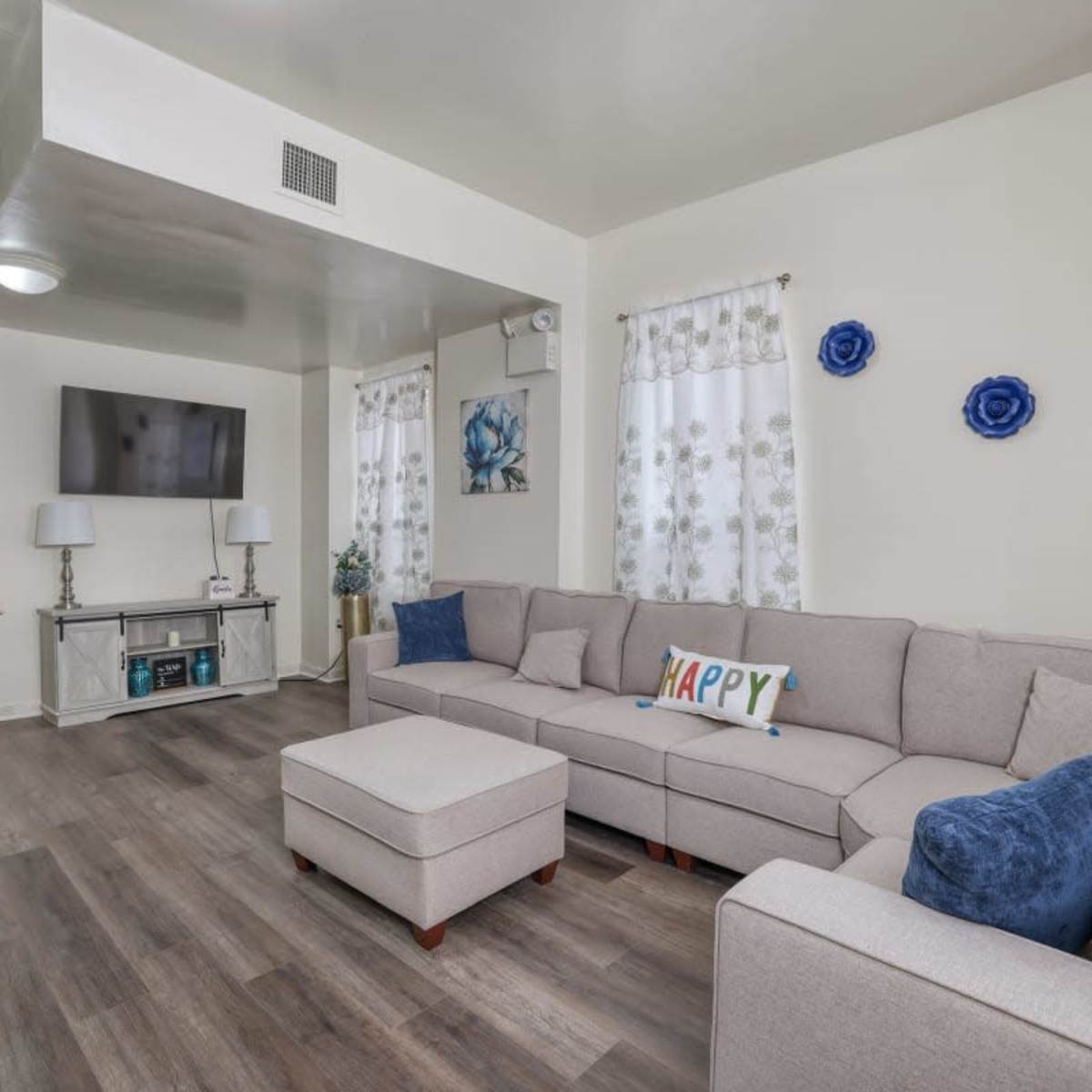 A modern living room with a gray sectional sofa, an ottoman, a television on the wall, and decorative plants.