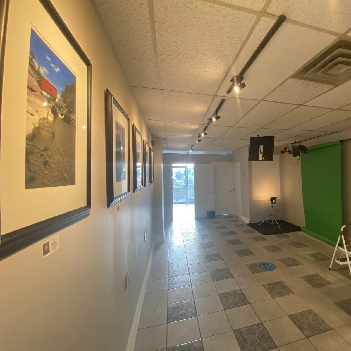 A hallway with framed photographs on the left, a green backdrop for video or photography at the far end, and a lighting setup on the right.