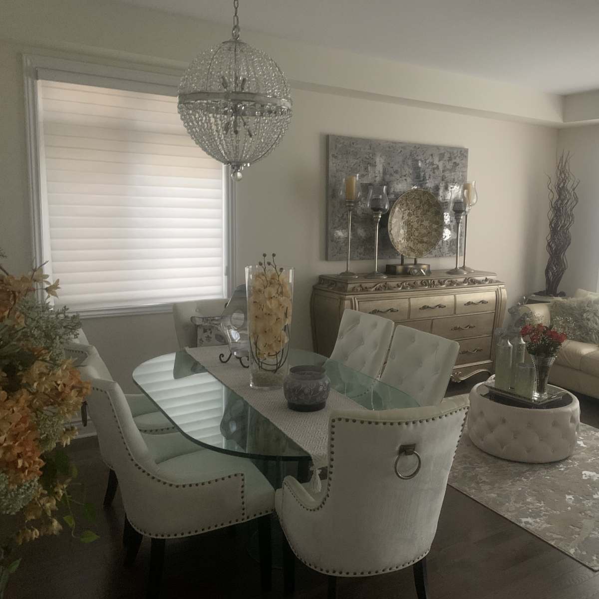 A modern living and dining area featuring a glass dining table with upholstered chairs, a cream-colored sofa, and decorative elements like a chandelier and a floral arrangement.