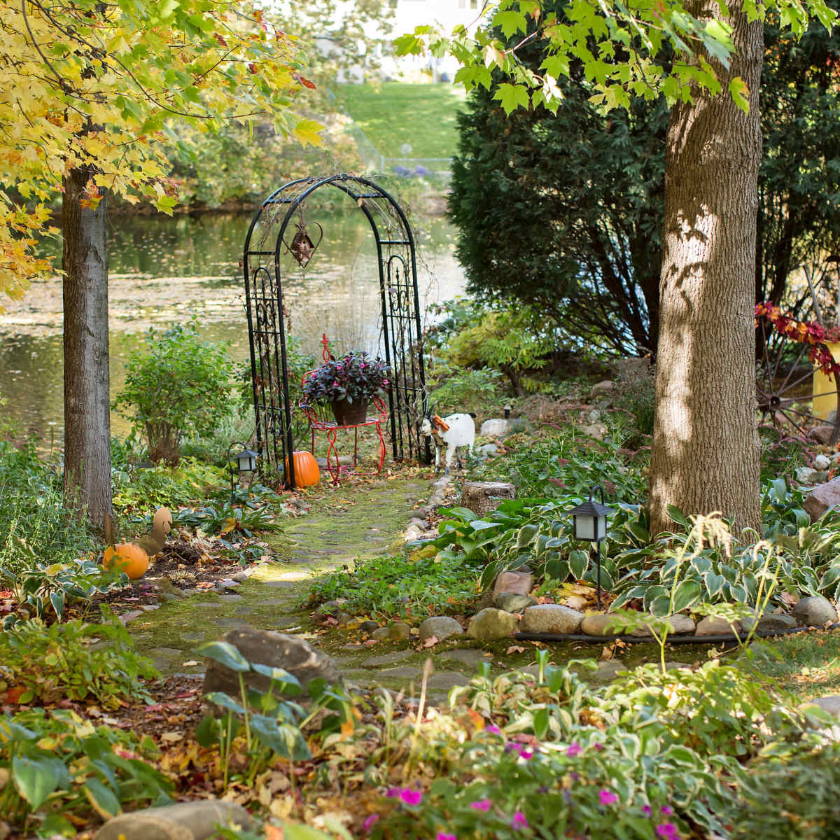 A stone path leads through a garden arch adorned with flowers, bordered by trees and a pond in the background.