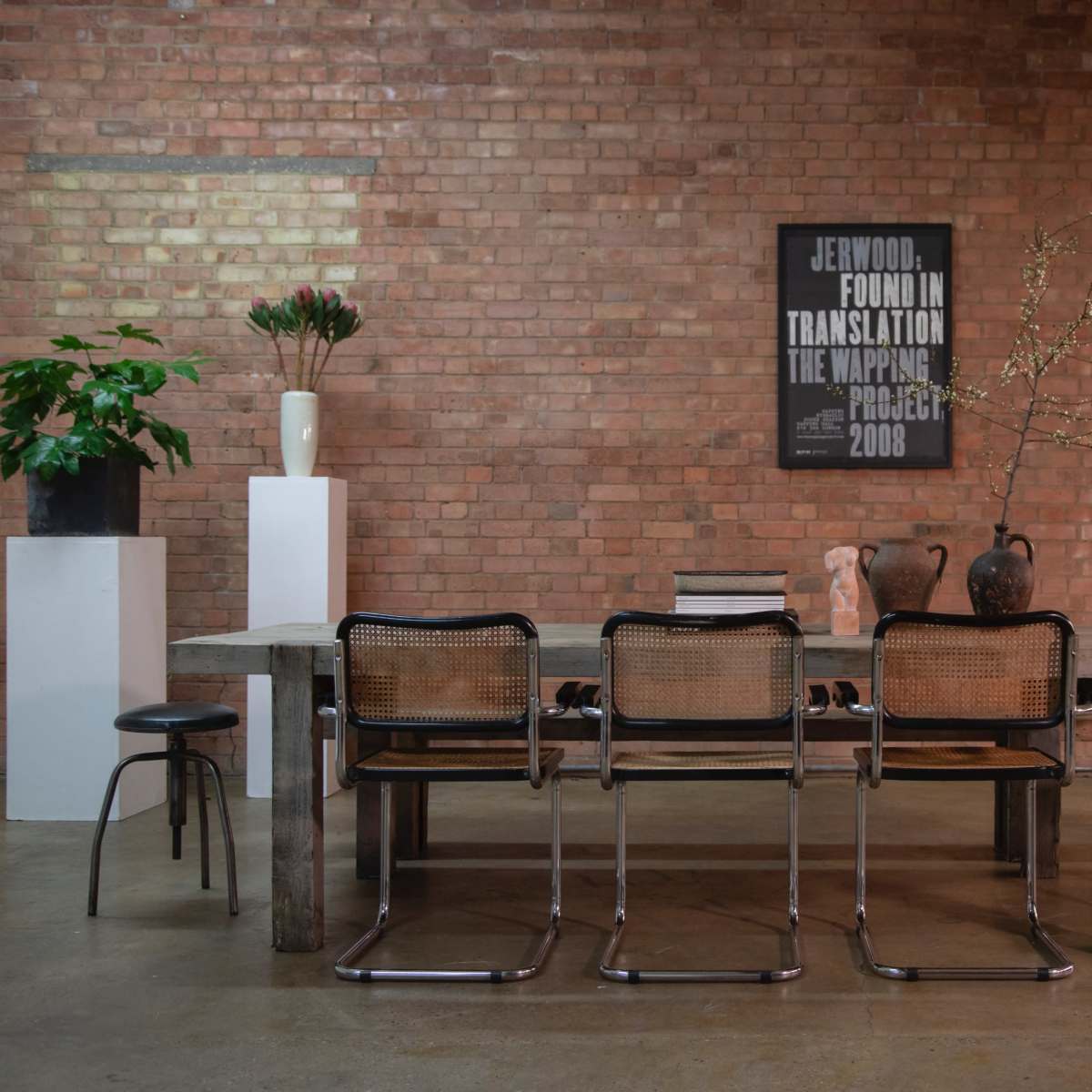 A wooden dining table is surrounded by metal chairs in an industrial-style room with exposed brick walls and various decorative plants and vases on display.