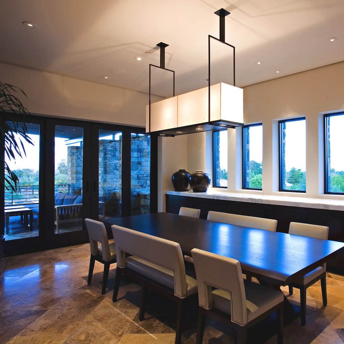 A spacious dining room features a long wooden table surrounded by upholstered chairs, with large windows allowing natural light and views of greenery.