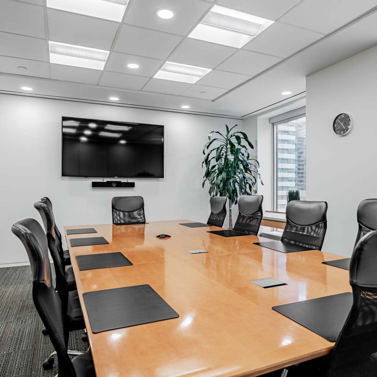 A modern conference room features a large wooden table surrounded by ergonomic black chairs, with a wall-mounted TV and a clock visible.