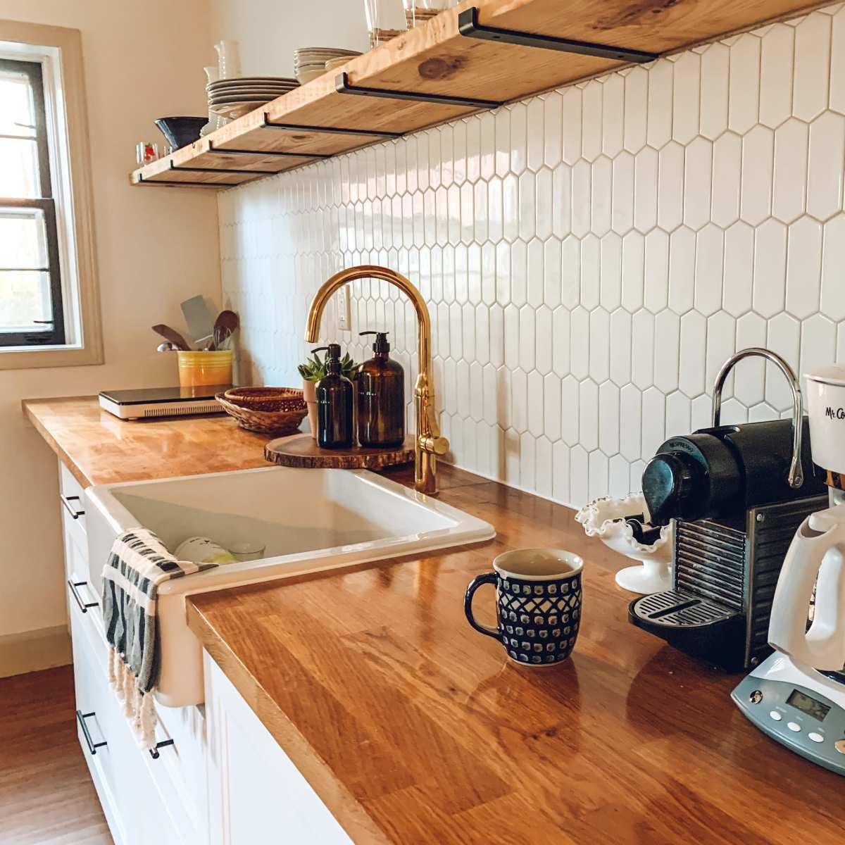 A kitchen countertop features a coffee maker, a kettle, and a mug next to a sink, with a backsplash of hexagonal white tiles and wooden shelves above displaying dishes.