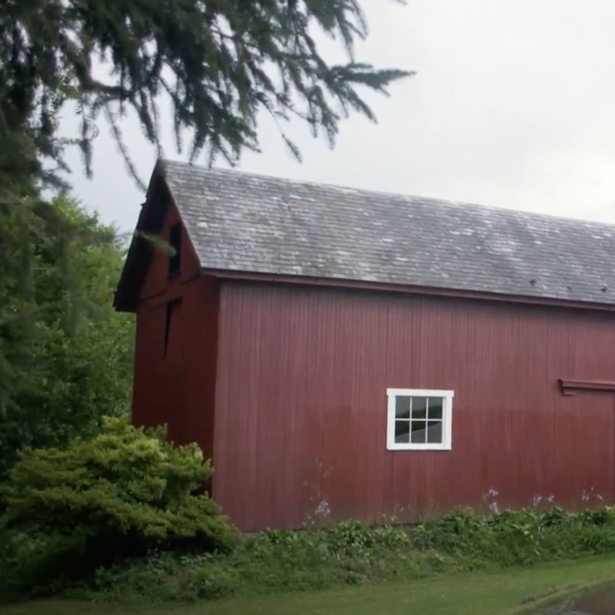 A red barn stands next to a white building, surrounded by green grass and trees under a cloudy sky.