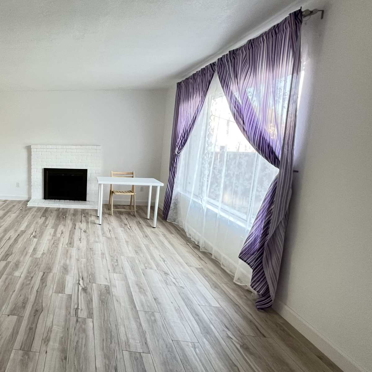 A spacious, unfurnished living room features a white fireplace, a small table with a chair, large windows with sheer curtains, and light-colored flooring.