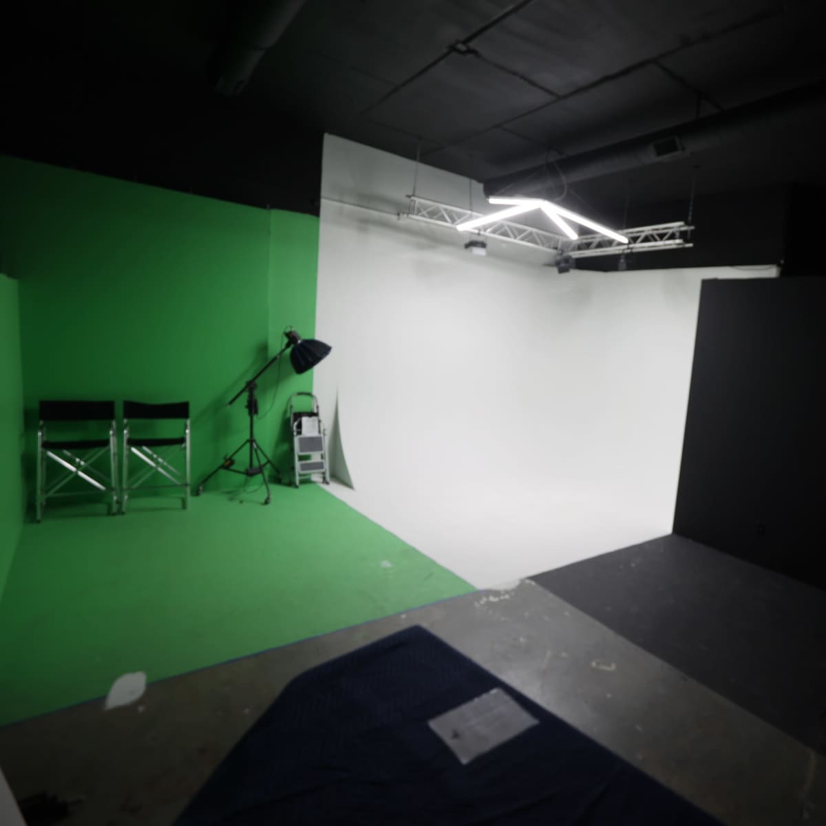 A photography studio featuring a green backdrop, a white backdrop, and a black wall, along with a ladder and a light fixture.