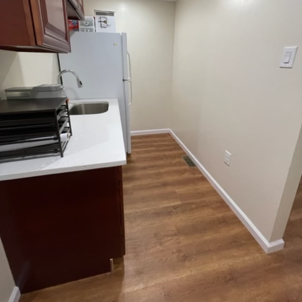 A narrow kitchen area with dark wooden cabinets, a sink, a white refrigerator, and light-colored walls and flooring.
