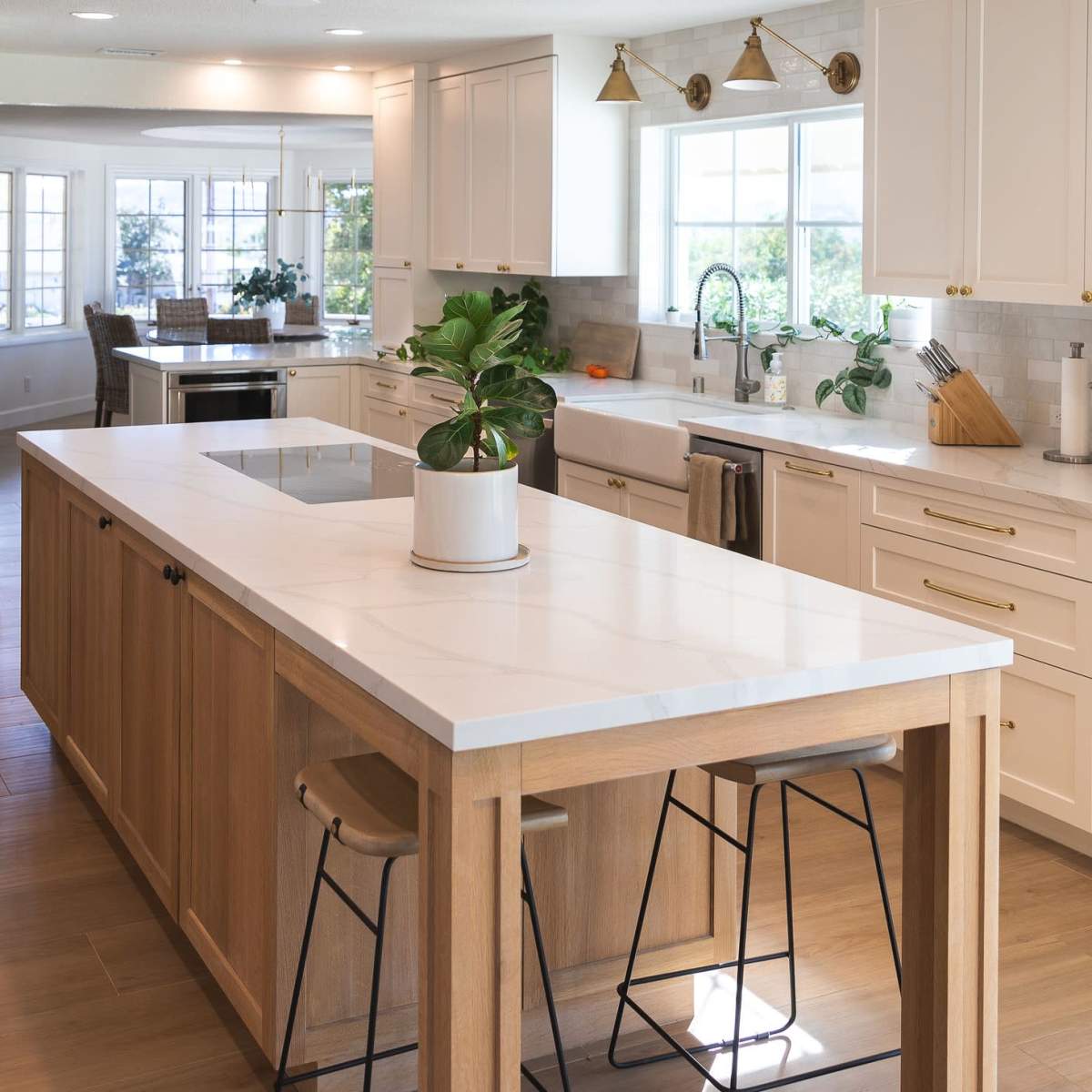 A modern kitchen features an island with a white countertop, wooden base, and bar stools, surrounded by cabinetry and large windows.