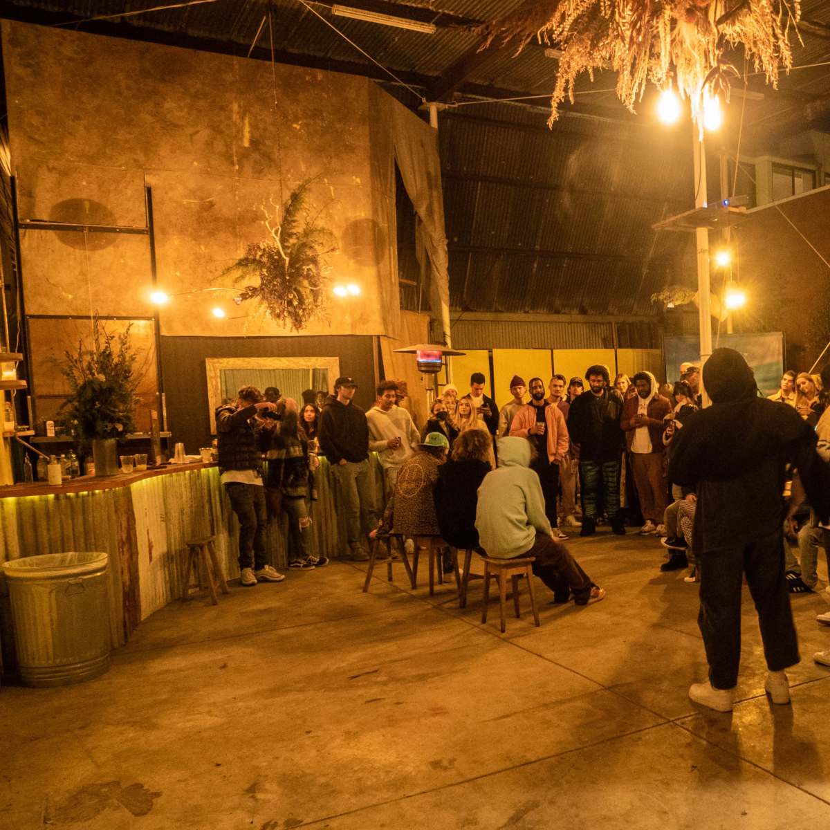 A crowd of people gathers in a dimly lit indoor space with rustic decor, wooden furniture, and hanging plants.