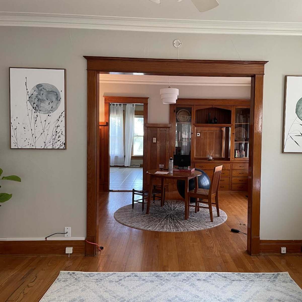 An interior setting with two framed artworks on a light gray wall, leading to a dining area furnished with a round table and four chairs.