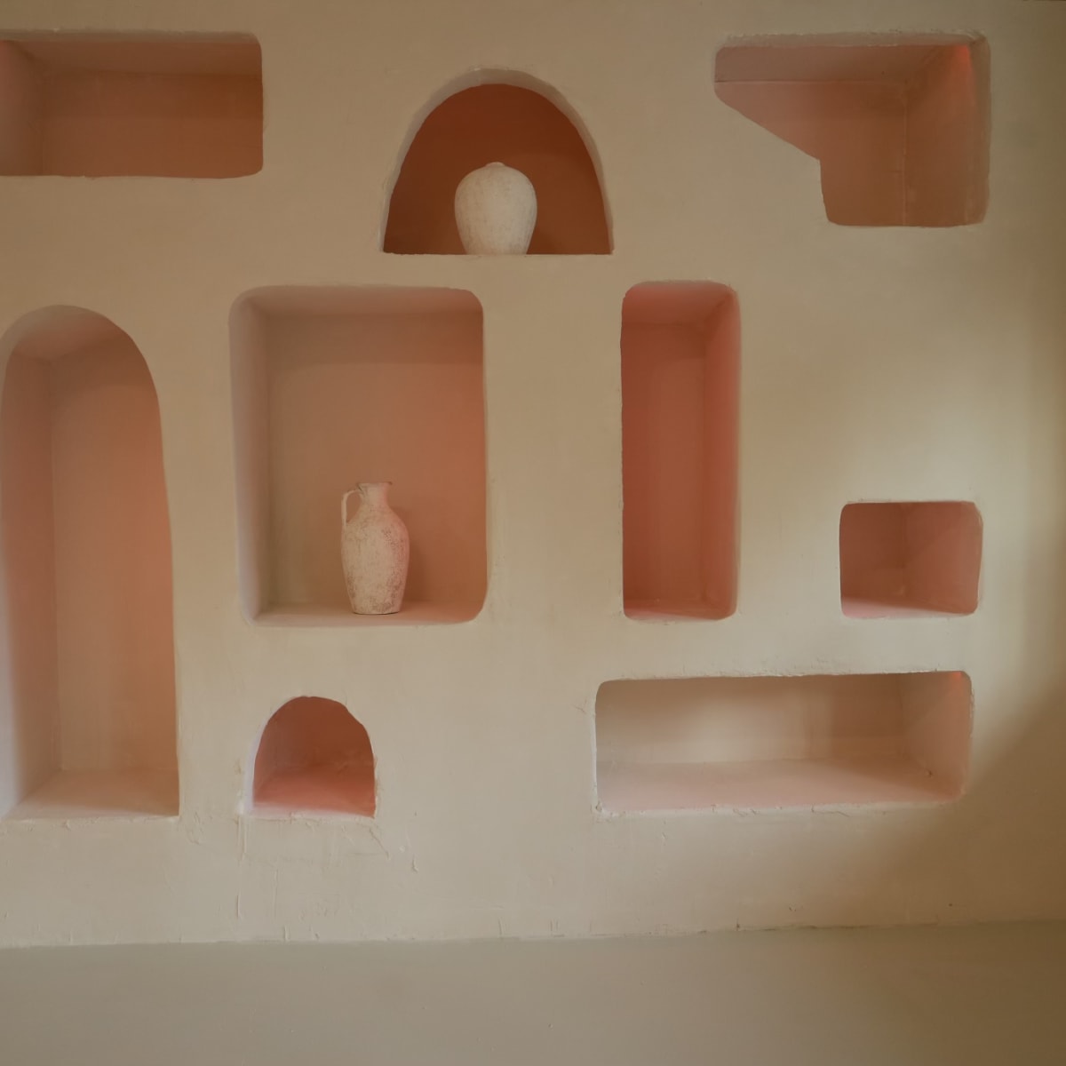 A textured wall displays several alcoves in various shapes, featuring a few decorative items including a vase and a pot.