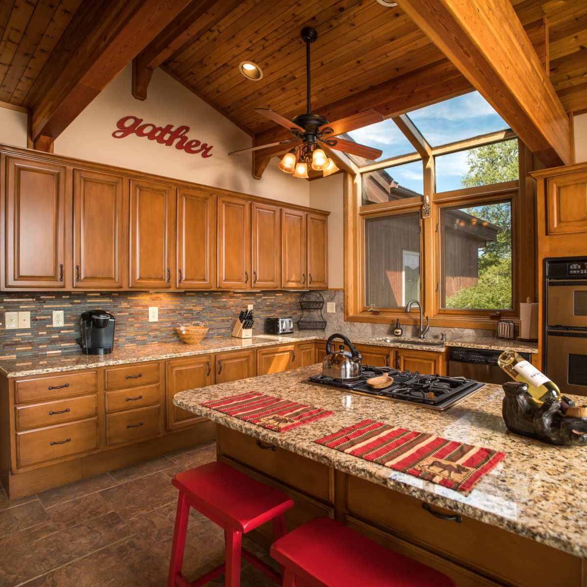 The kitchen features wooden cabinetry, a central island with red stools, and large windows that allow natural light to fill the space.