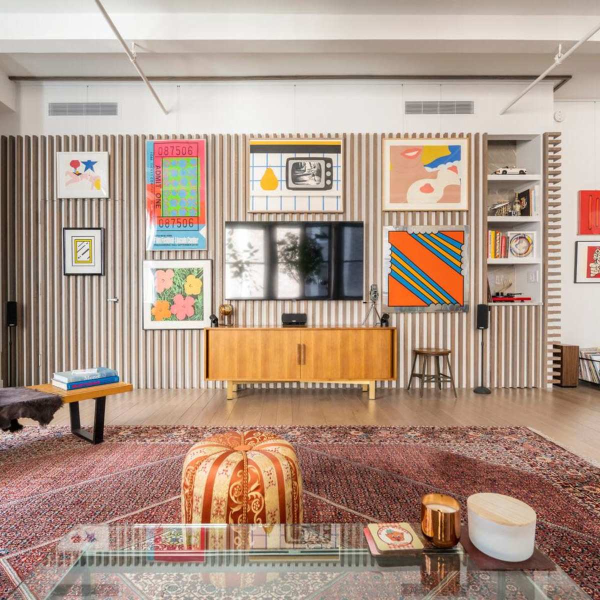 A modern living room features a wall of framed art and a wooden media console with a TV, surrounded by a patterned rug and various decorative items.
