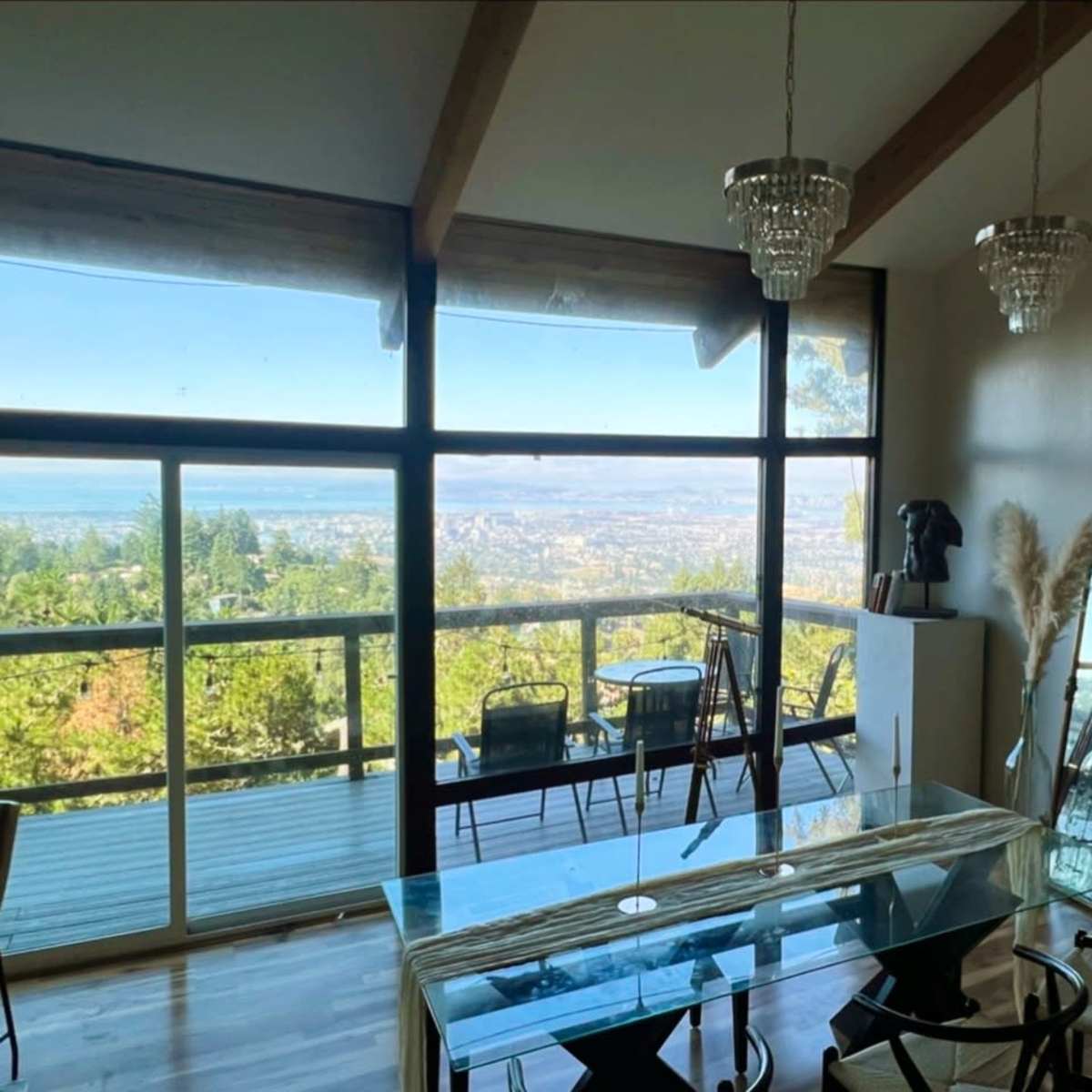 A dining area with a clear view of a landscape through large windows, featuring a glass table, black chairs, and decorative elements like mirrors and plants.