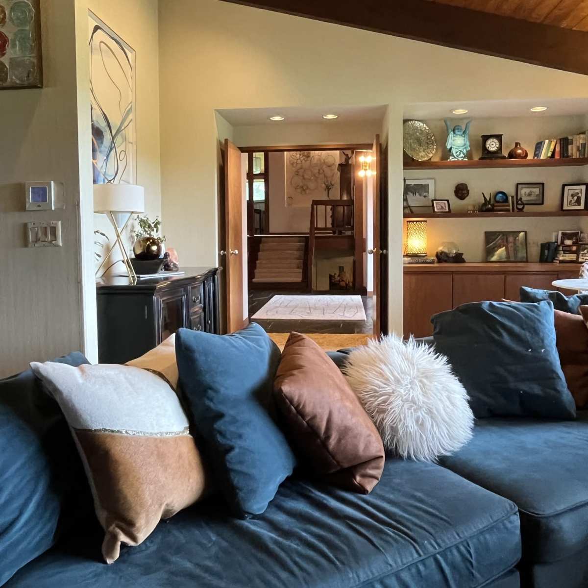 A living room features a blue couch adorned with decorative pillows and a view into a hallway leading to a staircase and an adjoining room.