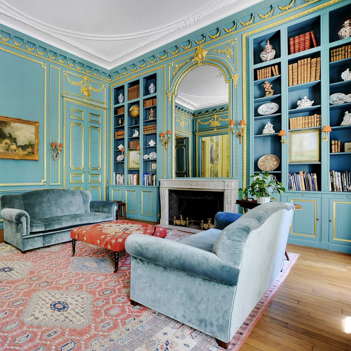 A spacious living room features light blue walls with ornate golden detailing, two green sofas, and a decorative bookshelf filled with books and artifacts.