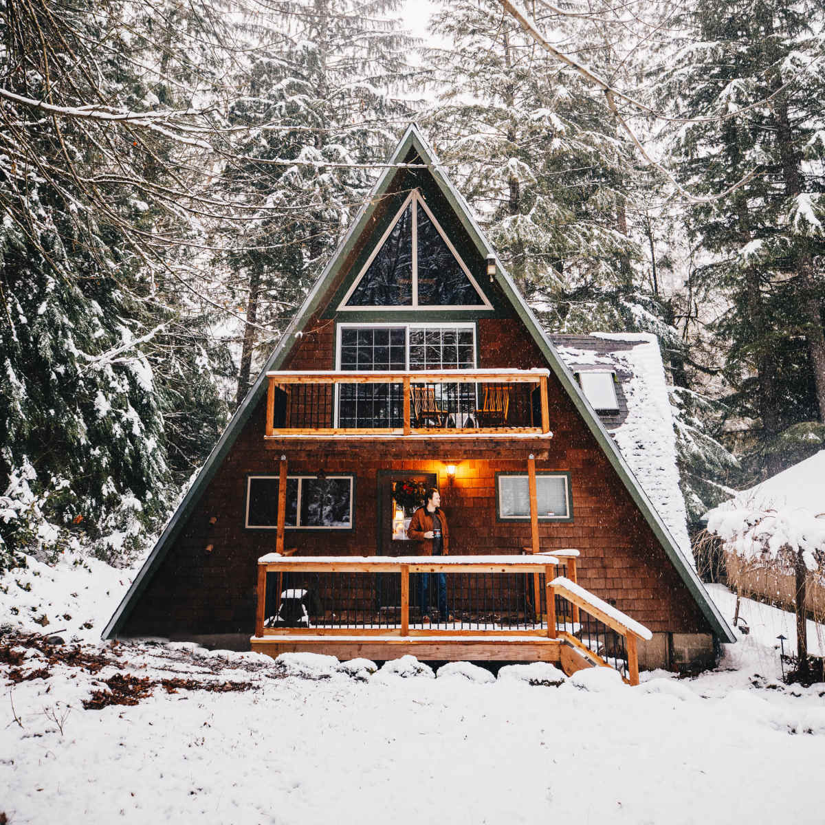 A wooden A-frame cabin with a large porch is surrounded by snow-covered trees in a winter landscape.