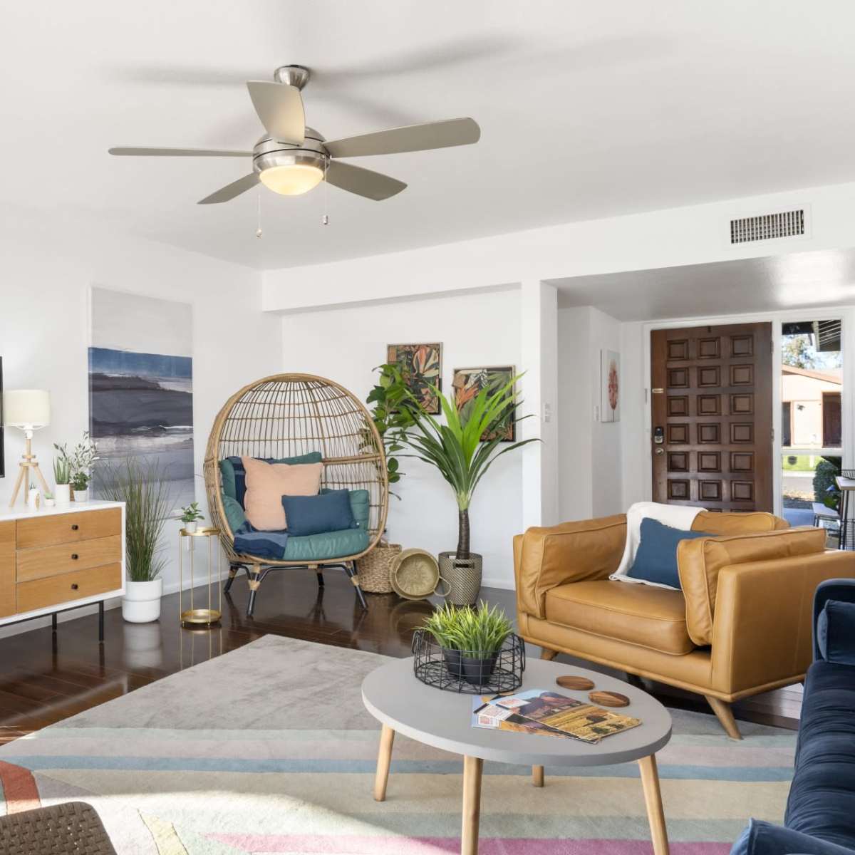 The living room features a television mounted on a white wall, a brown leather sofa, a circular coffee table, a decorative chair, and various plants.