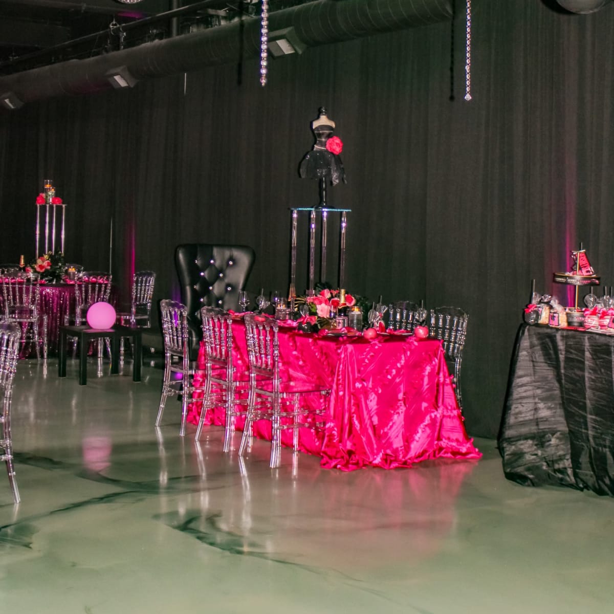 The scene features a finely decorated event space with black and pink balloon decorations, elegant tables covered in vibrant tablecloths, and chandeliers hanging from the ceiling.
