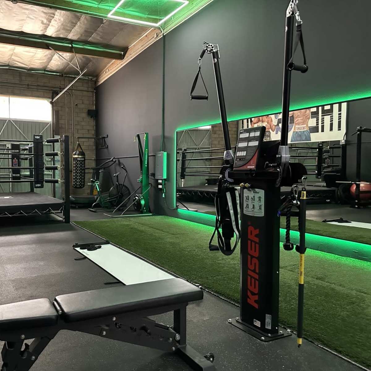 A modern gym interior featuring a weight bench, resistance training machine, boxing ring, and green turf area under bright lighting.