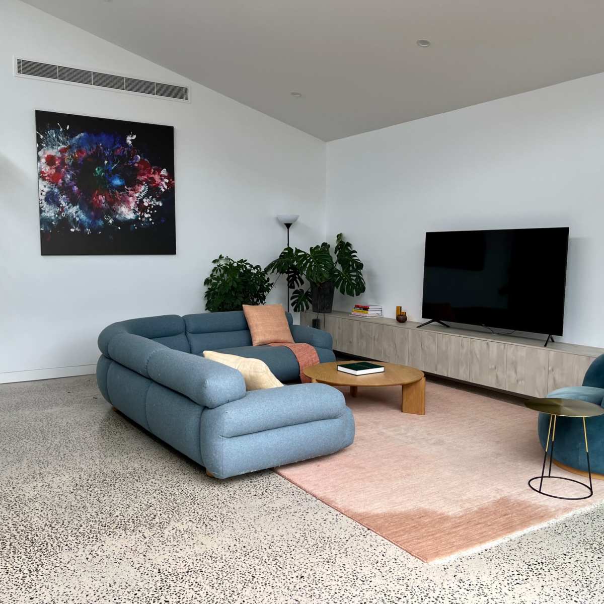 A modern living room features a blue curved sofa, a wooden coffee table, a television on a light-colored media unit, and green plants against white walls.