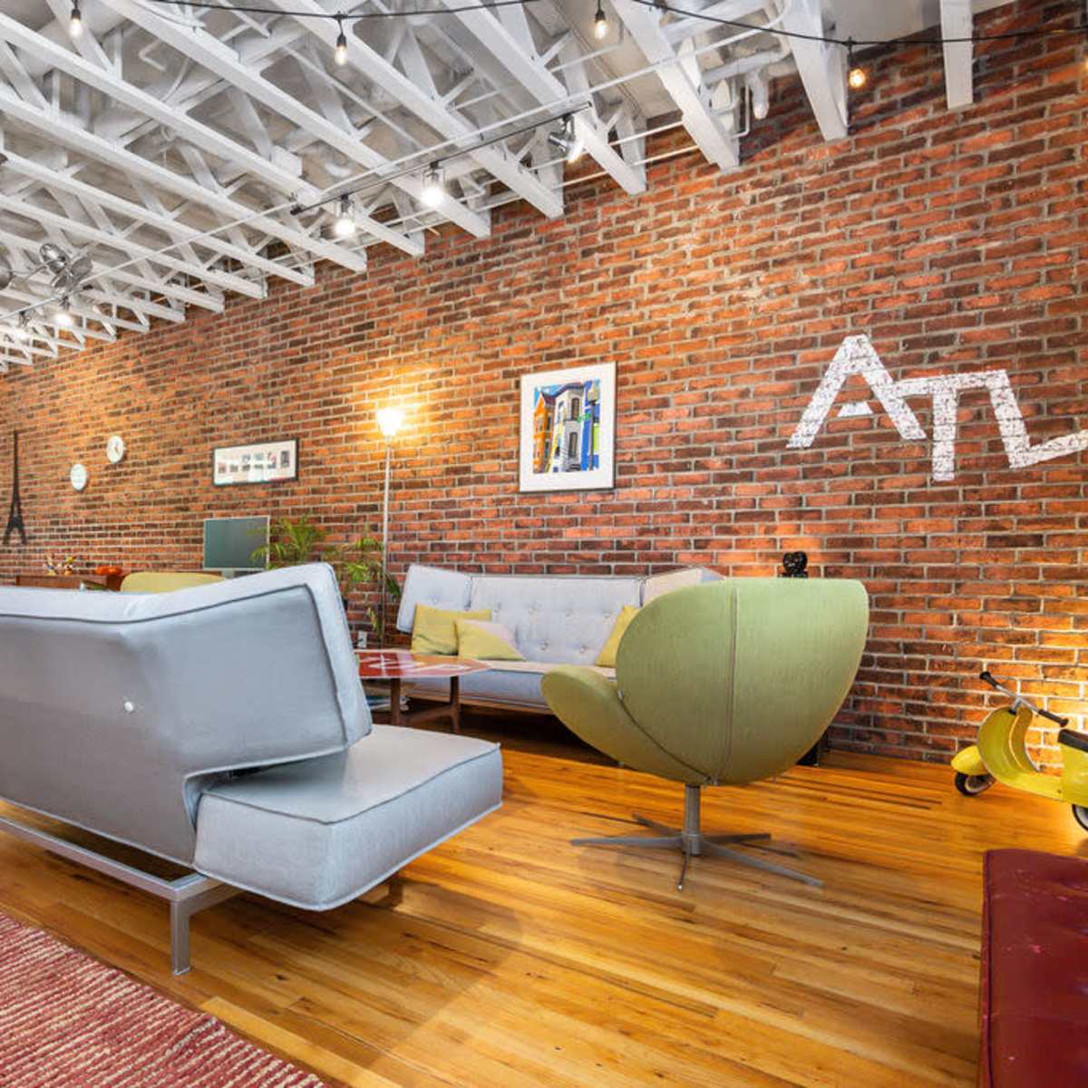 A modern living space with a combination of brick and wood elements, featuring colorful furniture and decorative artwork against a backdrop of exposed beams and overhead string lights.