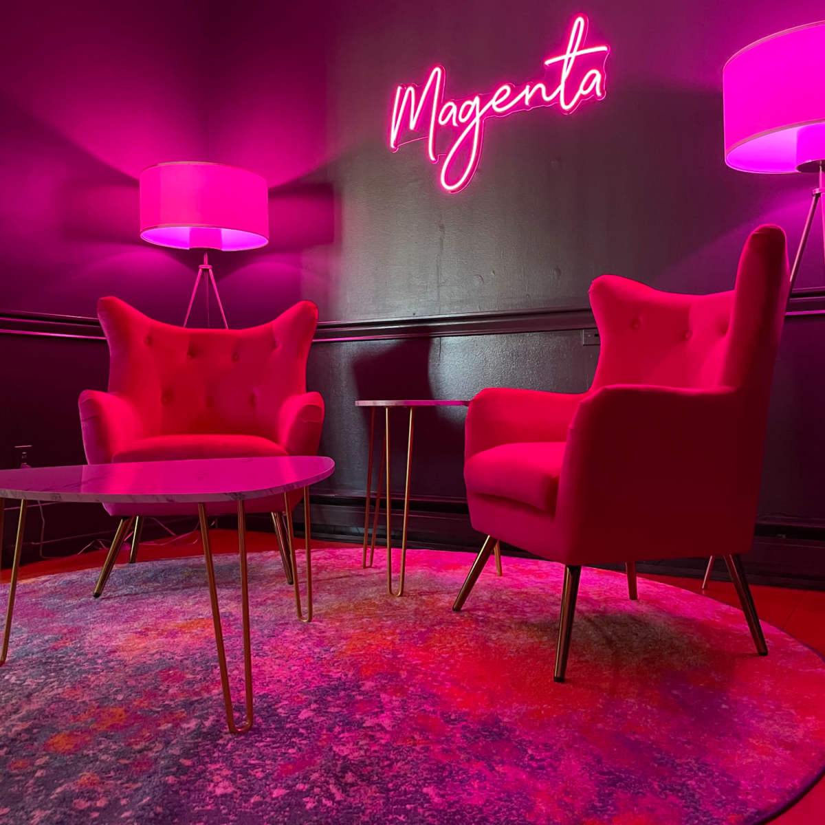 A room features two pink armchairs, a round table, and a brightly lit sign that spells "Magenta," all set against a dark wall.