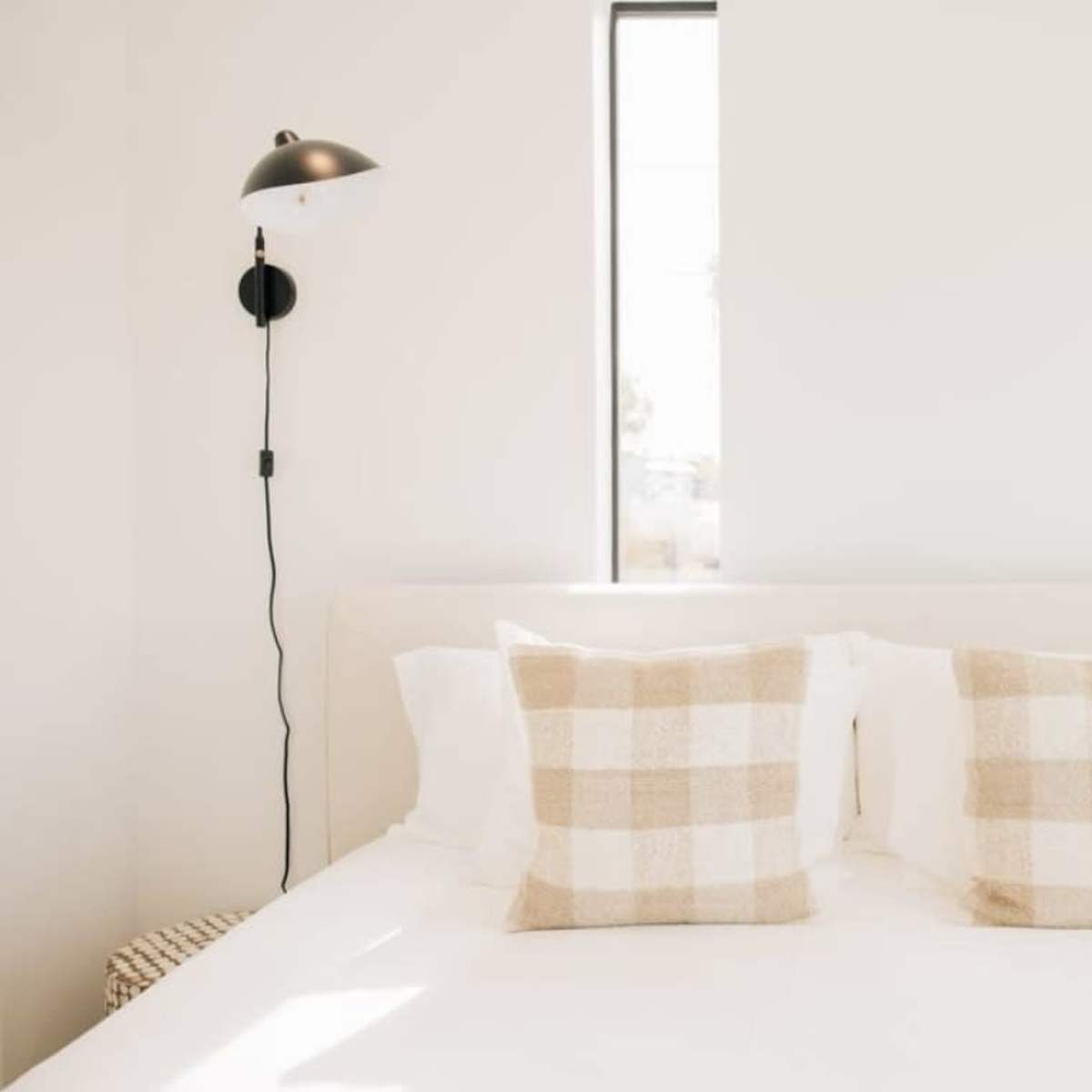 A bed with white bedding and two decorative pillows is positioned between wall-mounted lamps and a tall plant in a minimalist bedroom.