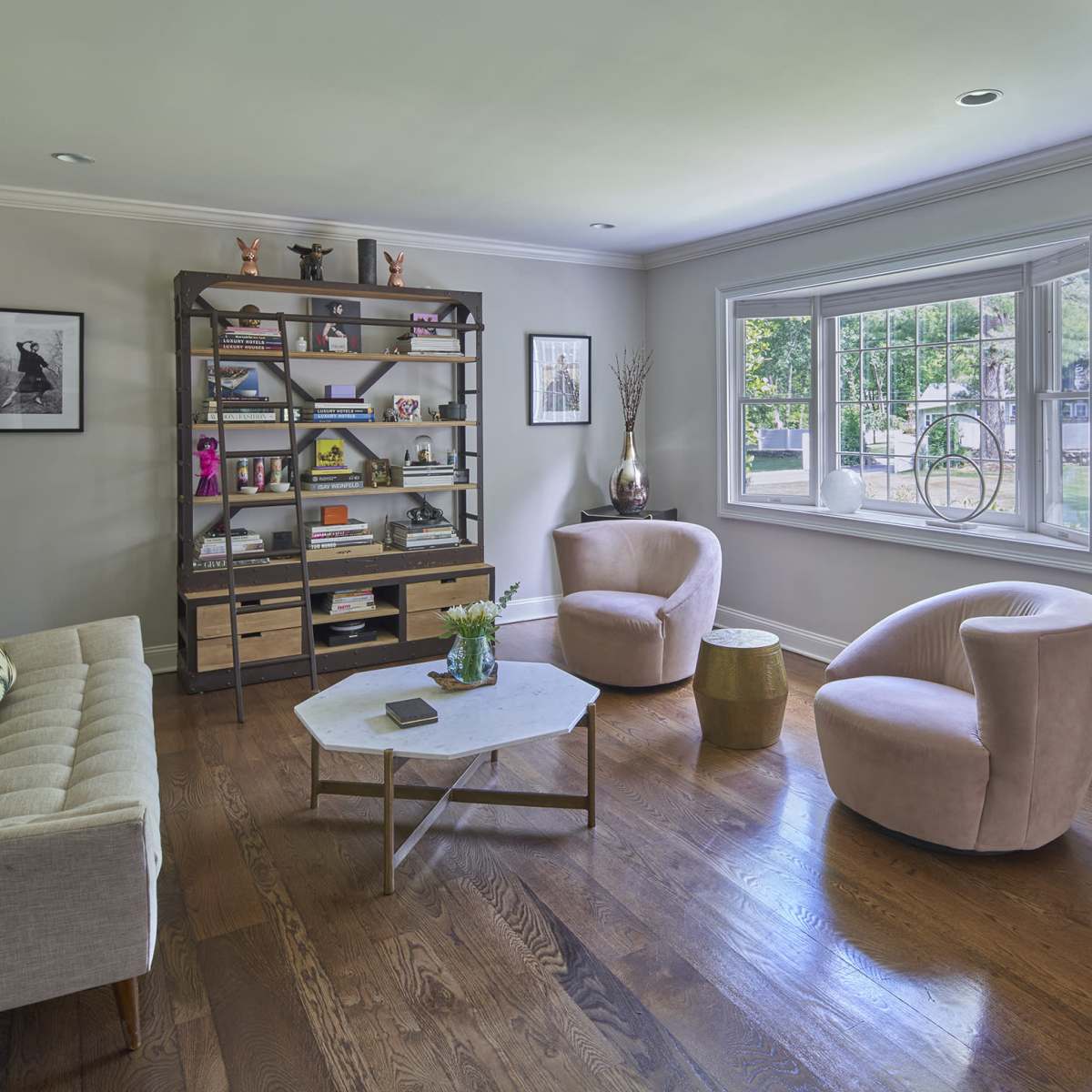 A modern living room features a gray sofa, two pink armchairs, a geometric coffee table, and a shelving unit filled with books and decorative items, with a window providing natural light.