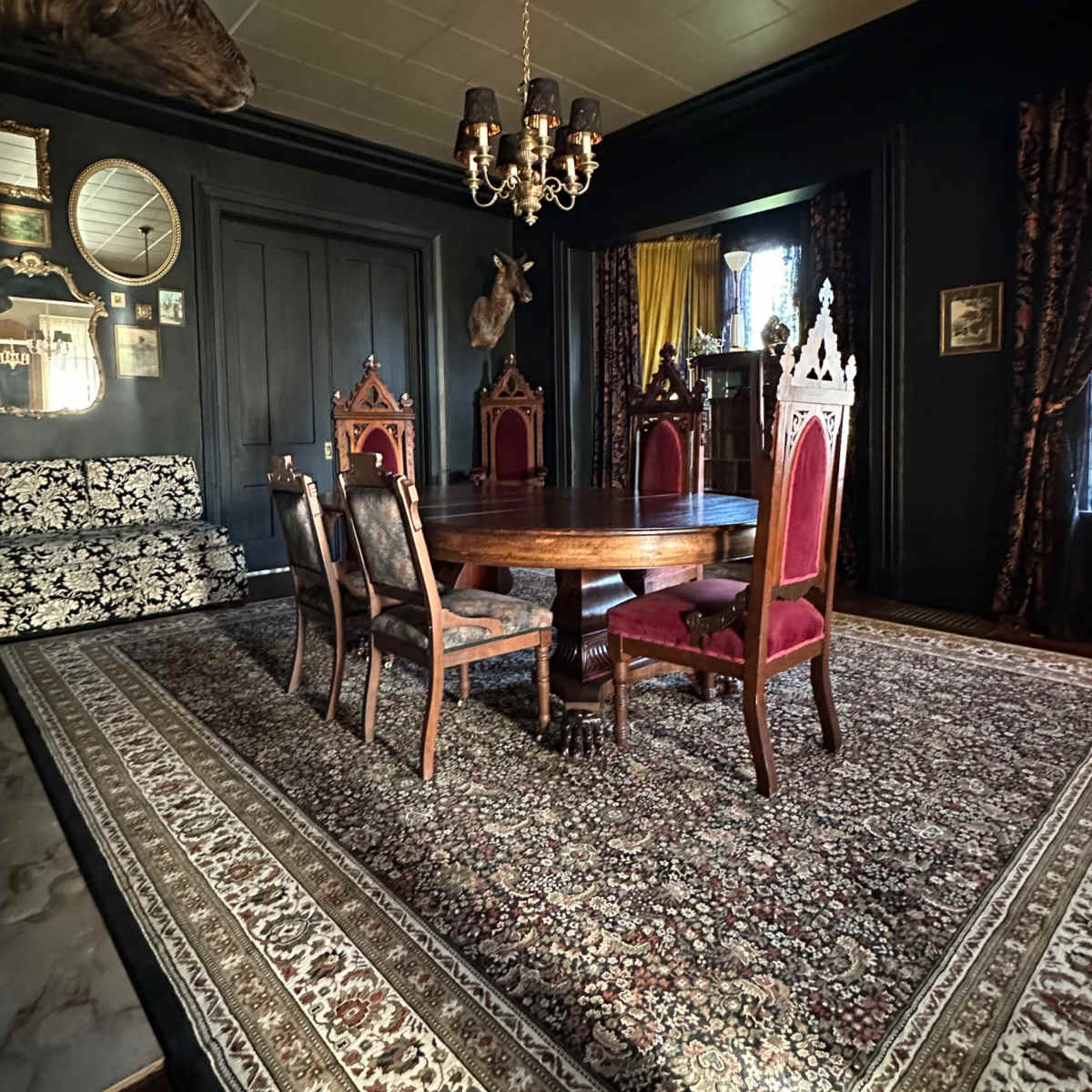 A darkly decorated dining room featuring a round wooden table surrounded by six chairs on a patterned rug, with ornate wall decorations and a chandelier overhead.