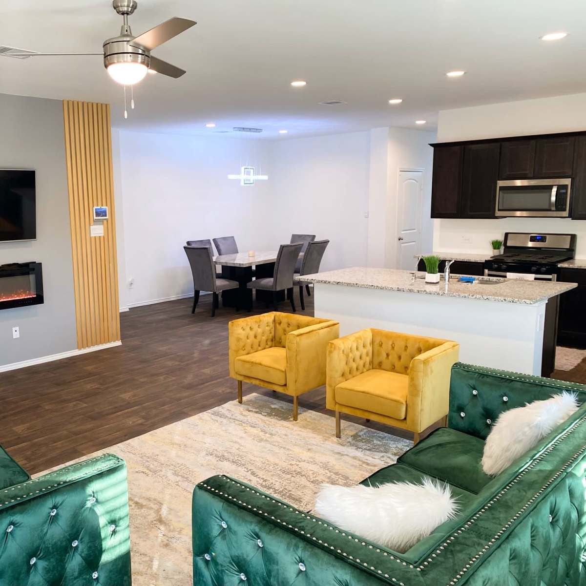 A modern living space featuring two green velvet sofas, a yellow armchair, a mounted television, an electric fireplace, and an adjacent dining area with a grey table and six chairs.